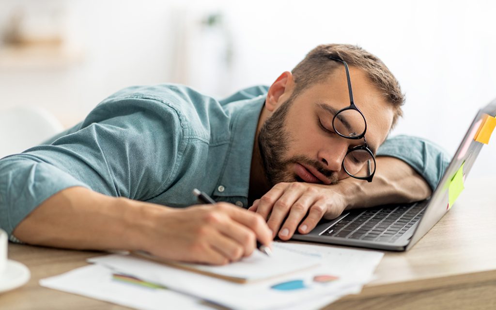 Exhausted millennial man sleeping on his office desk, next to laptop and documents, tired of overworking. Young male workaholic suffering from chronic fatigue at workplace